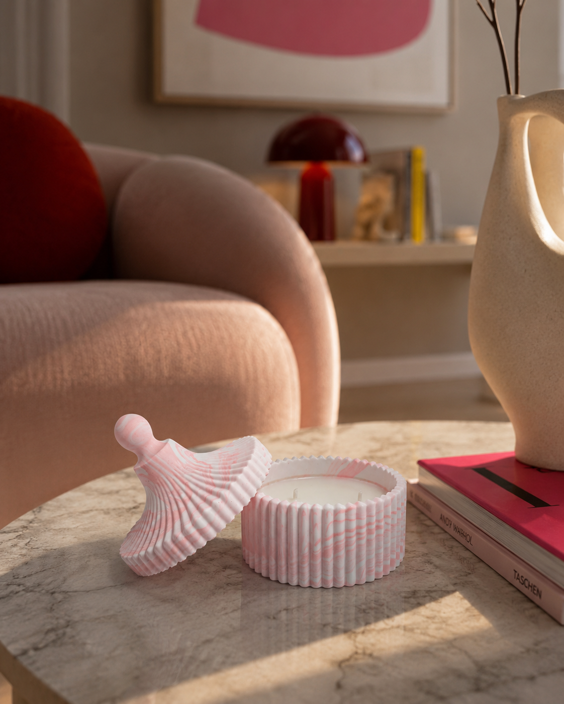 A pink and white striped candle in a ribbed round tin with its lid placed beside it on a marble table, set in a softly lit living room with a pink chair, books, and a vase in the background.